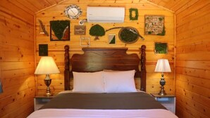 A wooden cabin bedroom with a bed, two bedside lamps, a wall-mounted air conditioner, and various wall decorations including framed pictures and a wreath.