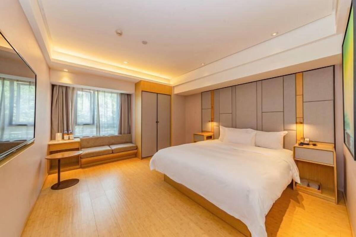 A modern hotel room with a large bed, bedside tables, a seating area, and a view of the city.
