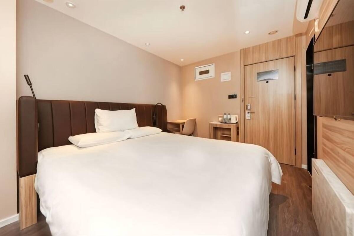 A hotel room with a large bed, a wooden wardrobe, a small table with a chair, and a desk with a chair.