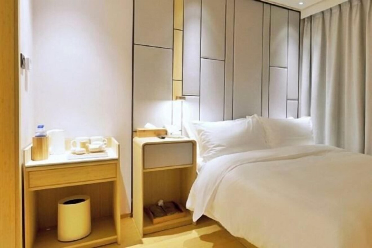 A hotel room with a bed, bedside table, and a small shelf with a trash bin.