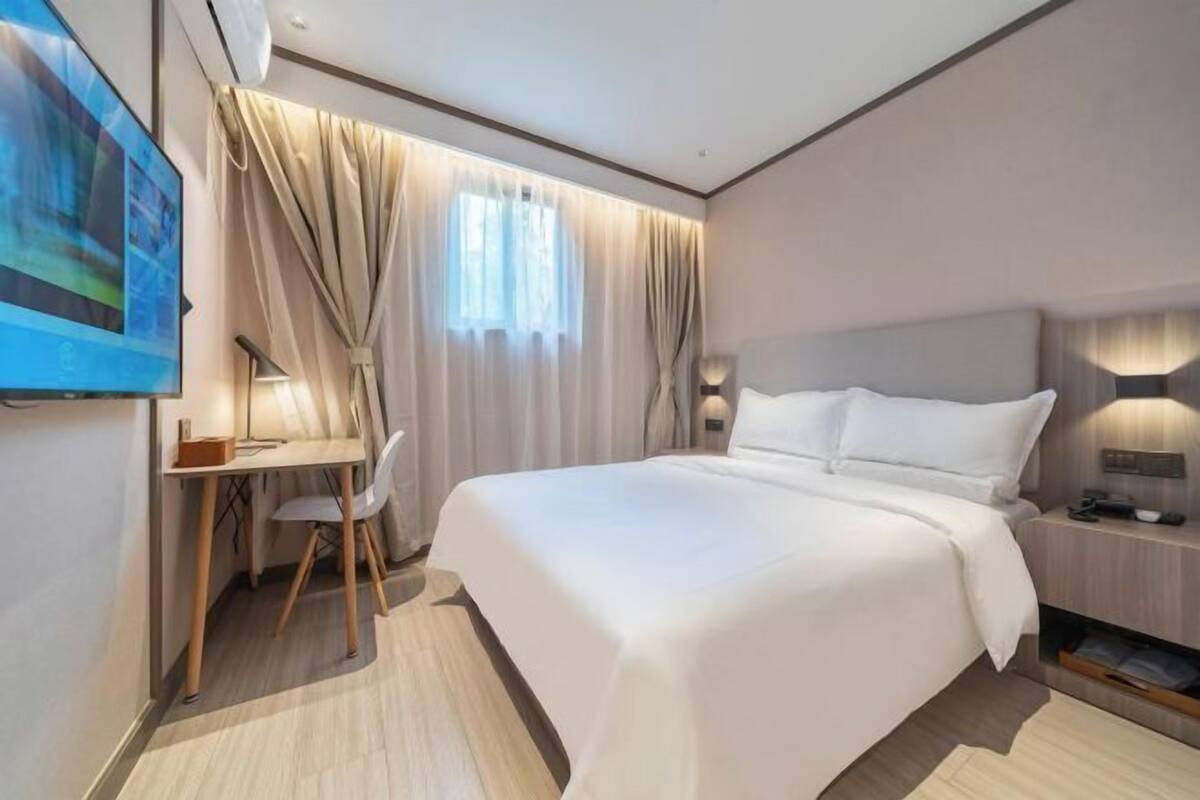 A modern hotel room with a large bed, a desk with a chair, a TV, and a nightstand with a lamp.
