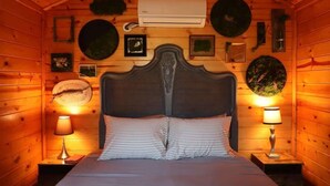 Una habitación tipo cabaña de madera, con cama, dos mesitas de noche, aire acondicionado instalado en la pared y diversas decoraciones.