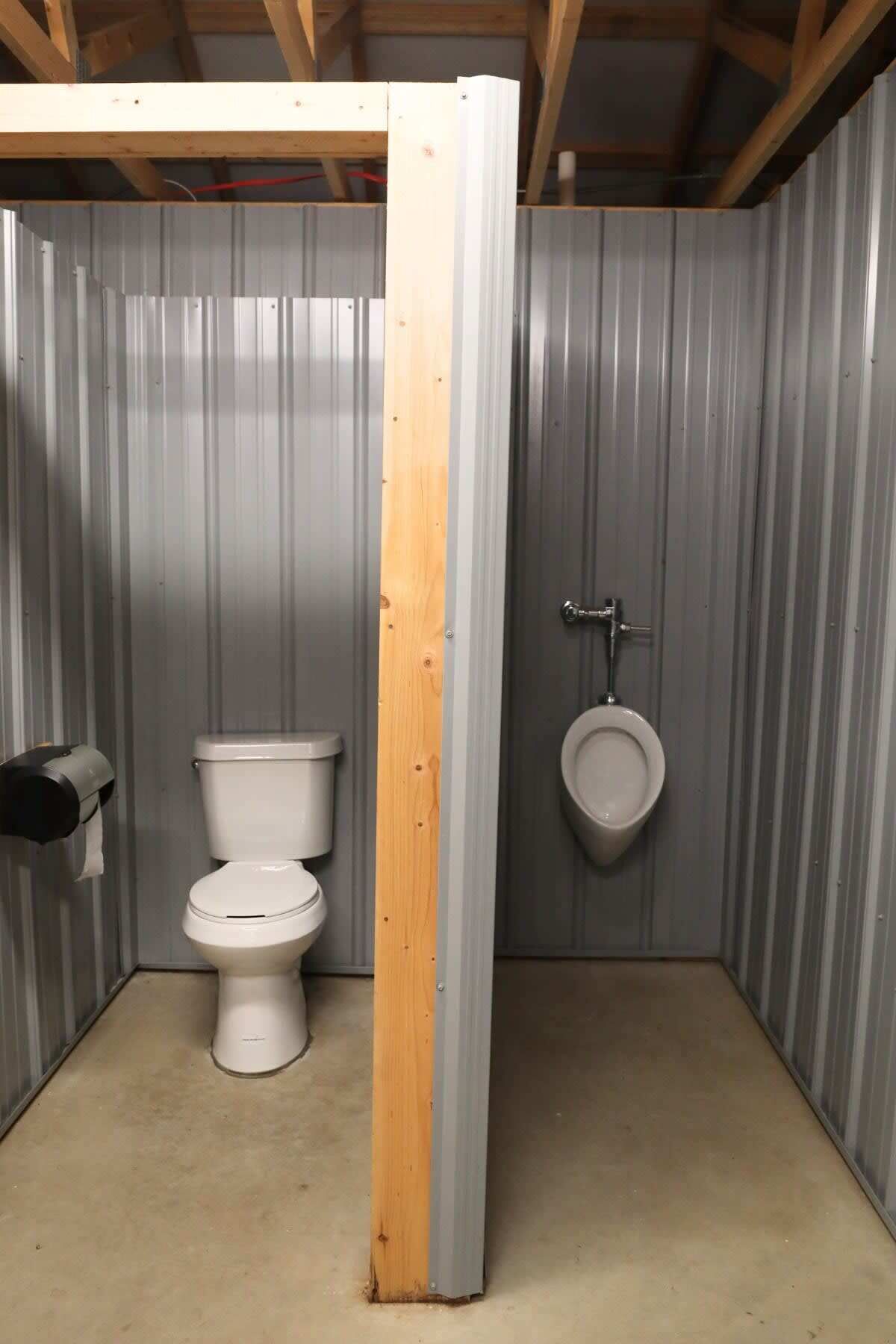 Un baño pequeño y cerrado con inodoro y orinal, paredes de metal y una viga de soporte de madera.