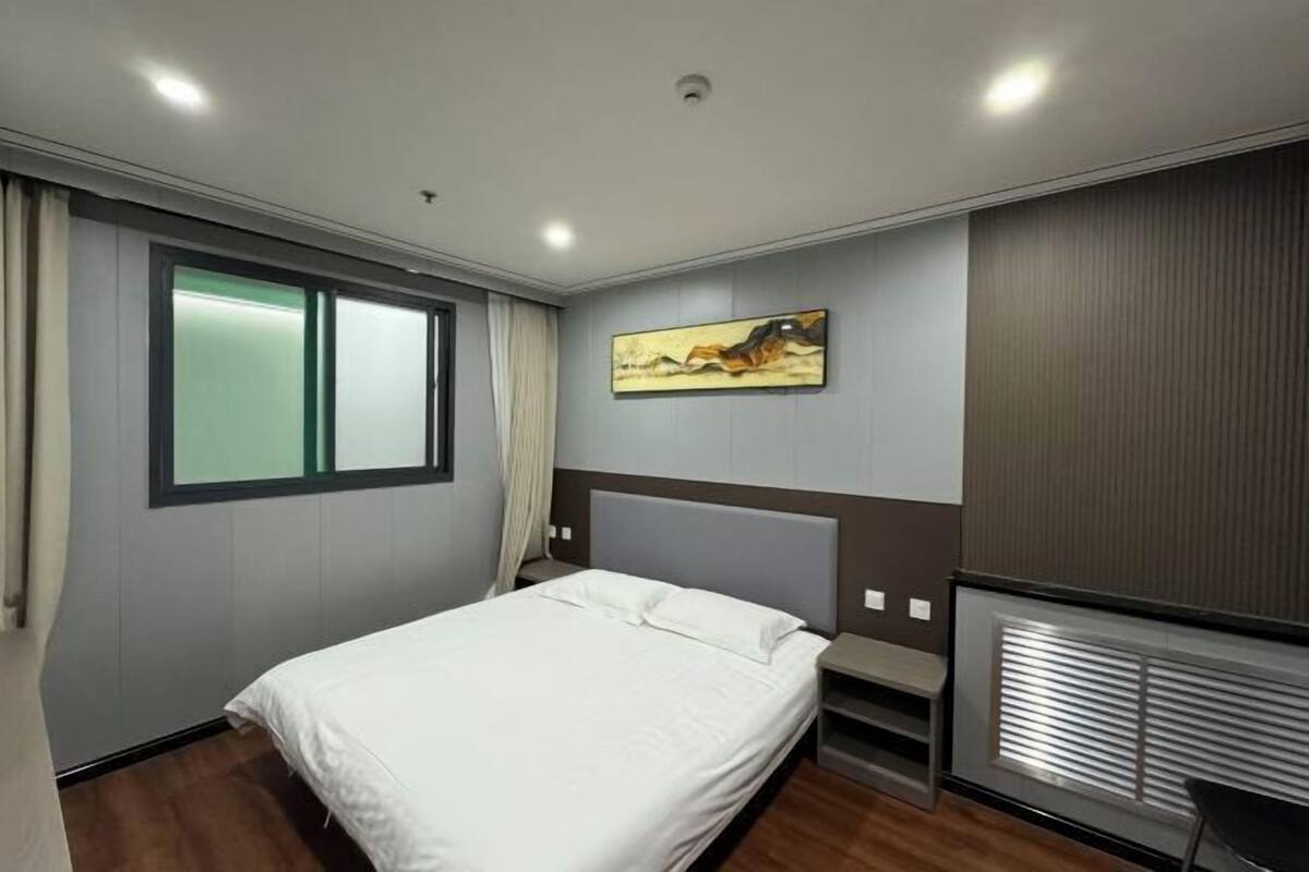 A hotel room with a bed, a window with curtains, a painting on the wall, and a built-in heater.