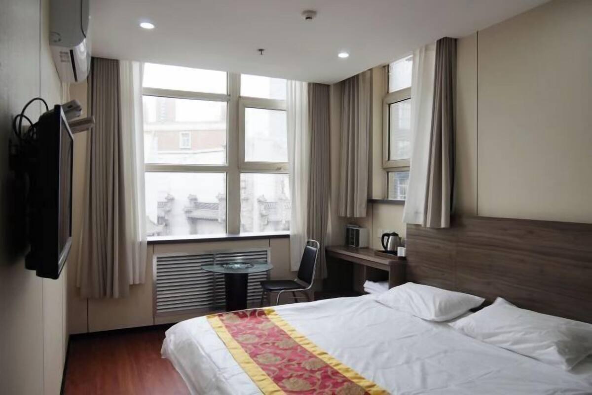 A hotel room with a bed, a desk, a chair, a window with curtains, and a view of buildings outside.
