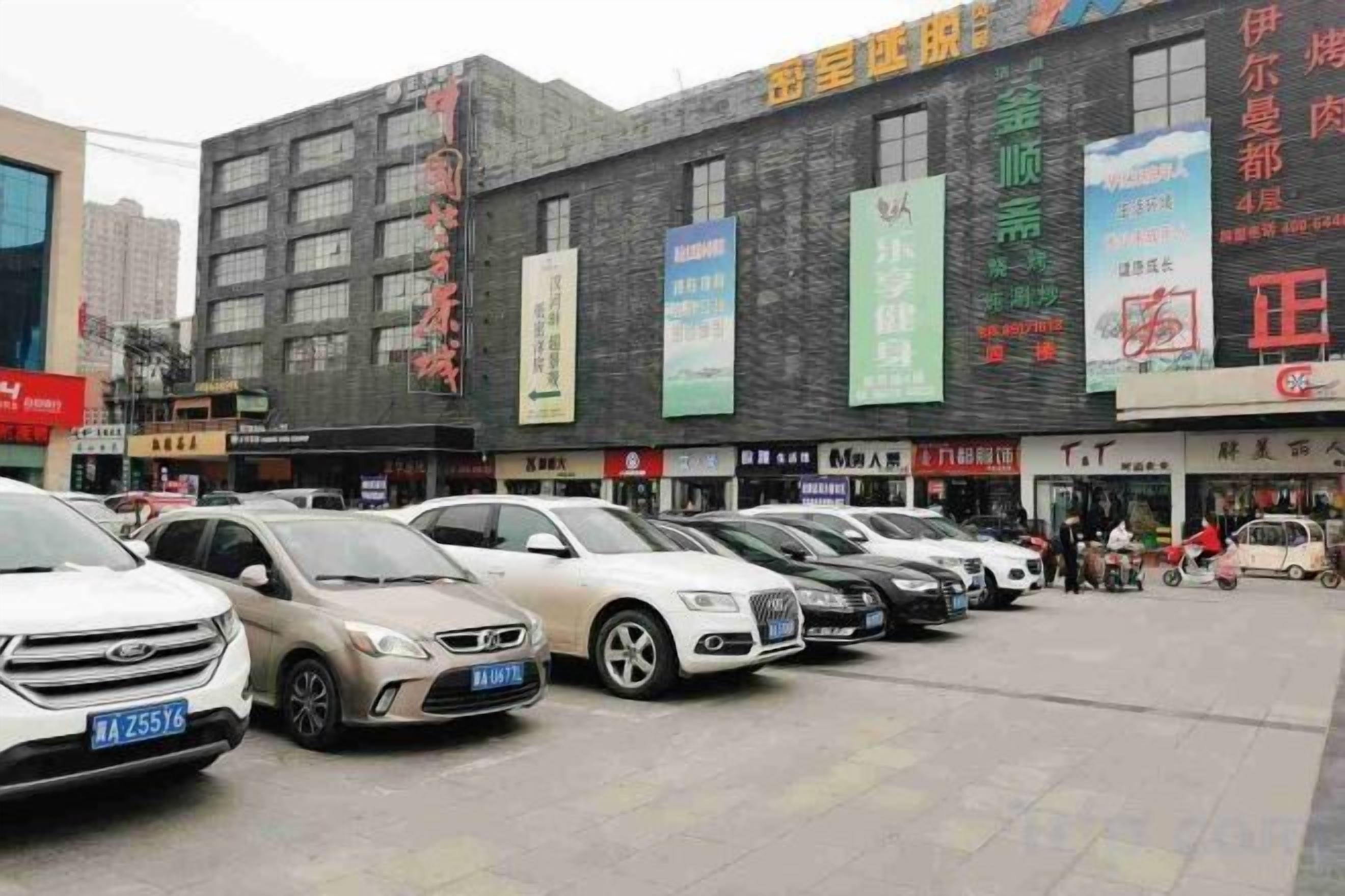 Ein Parkplatz mit mehreren Autos, ein Gebäude mit verschiedenen Schildern und einige Personen im Hintergrund.