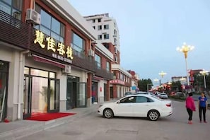 A street view with a building featuring a red carpet entrance, a white car parked in front, and a few pedestrians. - Home Inn Huayi Hotel (Changdao Scenic Area) (Yantai)