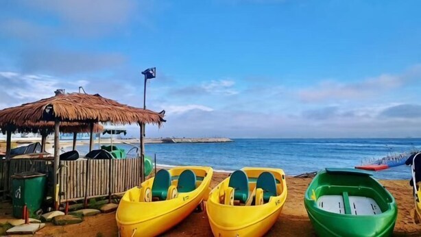 Una platja amb una cabana de palla, una filera de kayak grocs i verds, i un cel blau clar.
