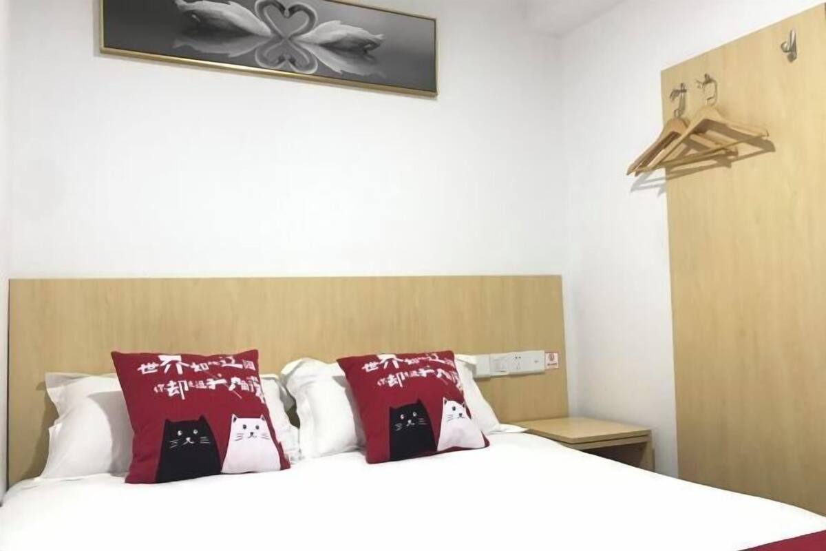 A hotel room with a bed, two pillows with cat designs, a wooden headboard, a wall-mounted light switch, and a wooden wardrobe with a hanger.