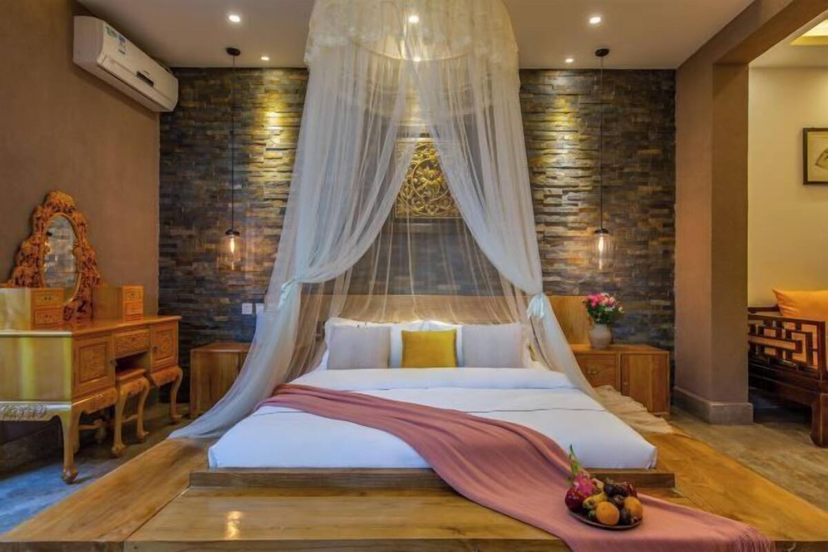 Een slaapkamer met een bed, nachtkastje, houten dressoir, decoratieve spiegel en een stenen muur.