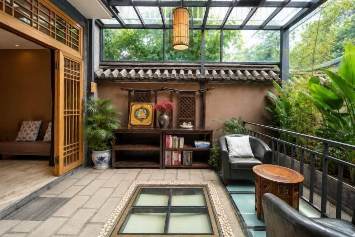 Een kamer in traditionele stijl met een tatami mat, een houten plank met boeken en decoratieve objecten, een comfortabele zithoek en uitzicht op groen door een glazen dak.