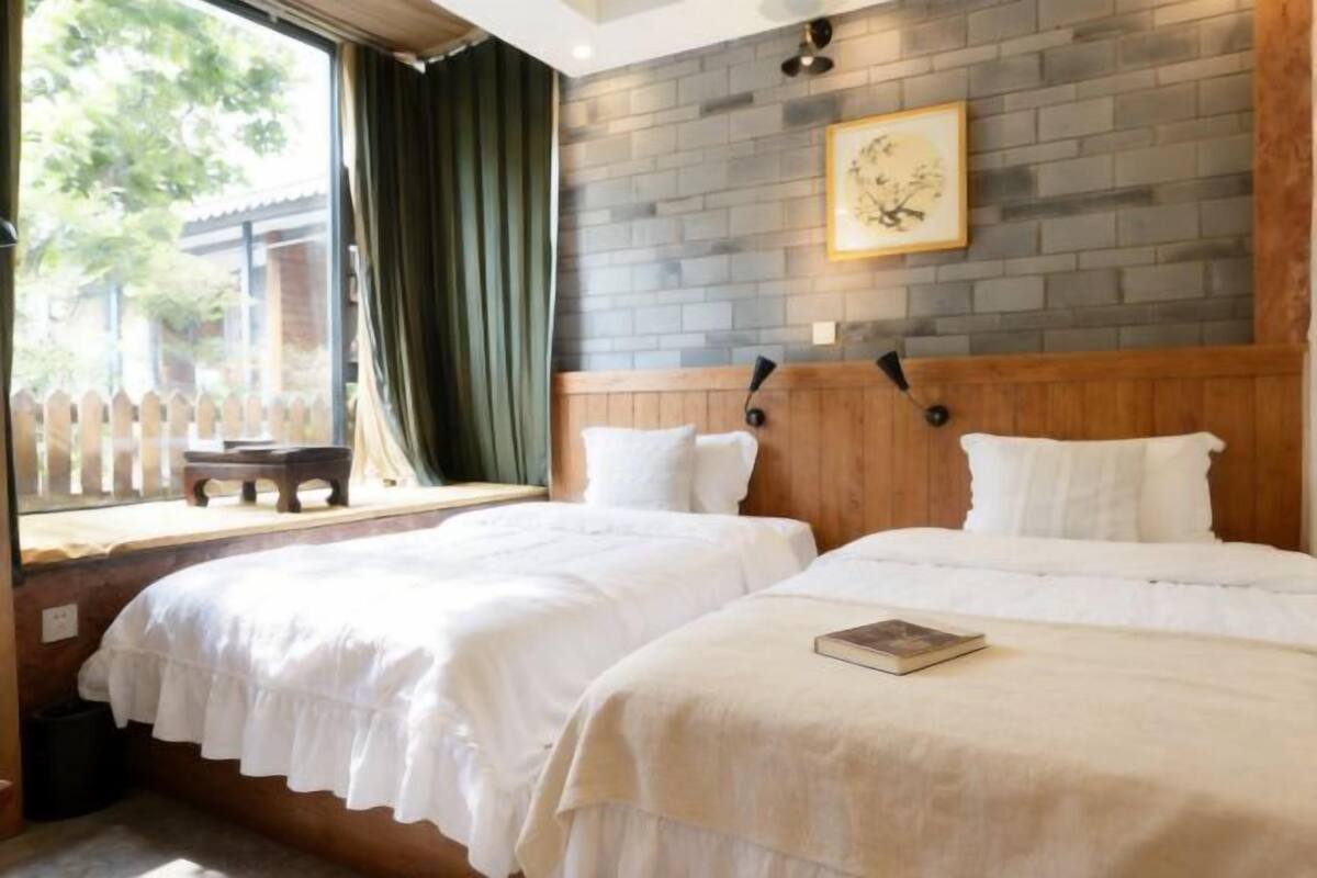 Een kamer met twee bedden, een houten hoofdbord, een bakstenen muur en een raam met uitzicht op groen.