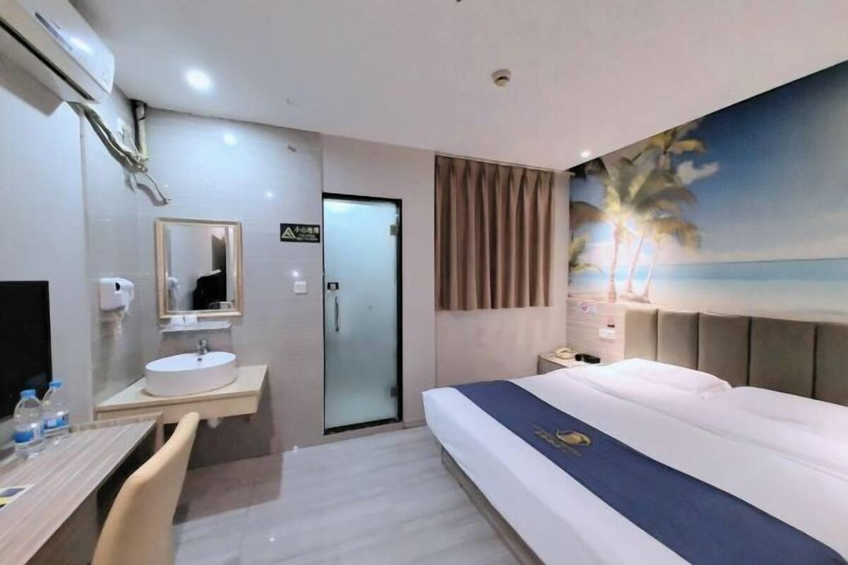A hotel room with a bed, a bedside table, a desk with a chair, a bathroom with a sink and mirror, and a painting of a beach scene.