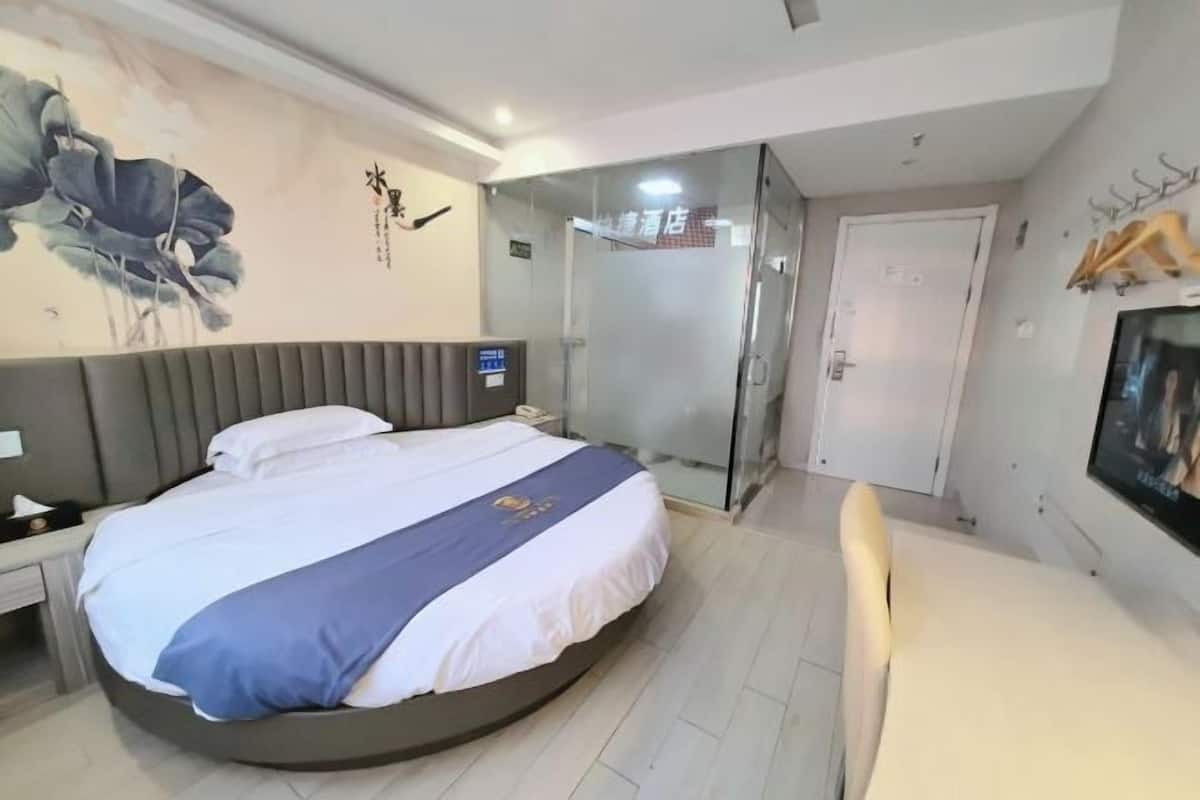 A hotel room with a round bed, a glass shower enclosure, a television, and a wall with a floral painting.