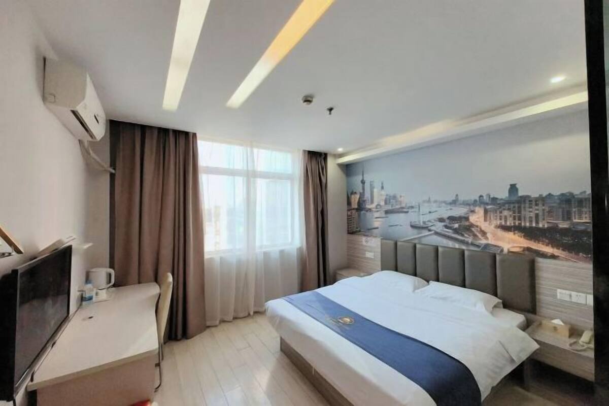 A hotel room with a bed, a desk with a TV, a window with curtains, and a cityscape mural on the wall.