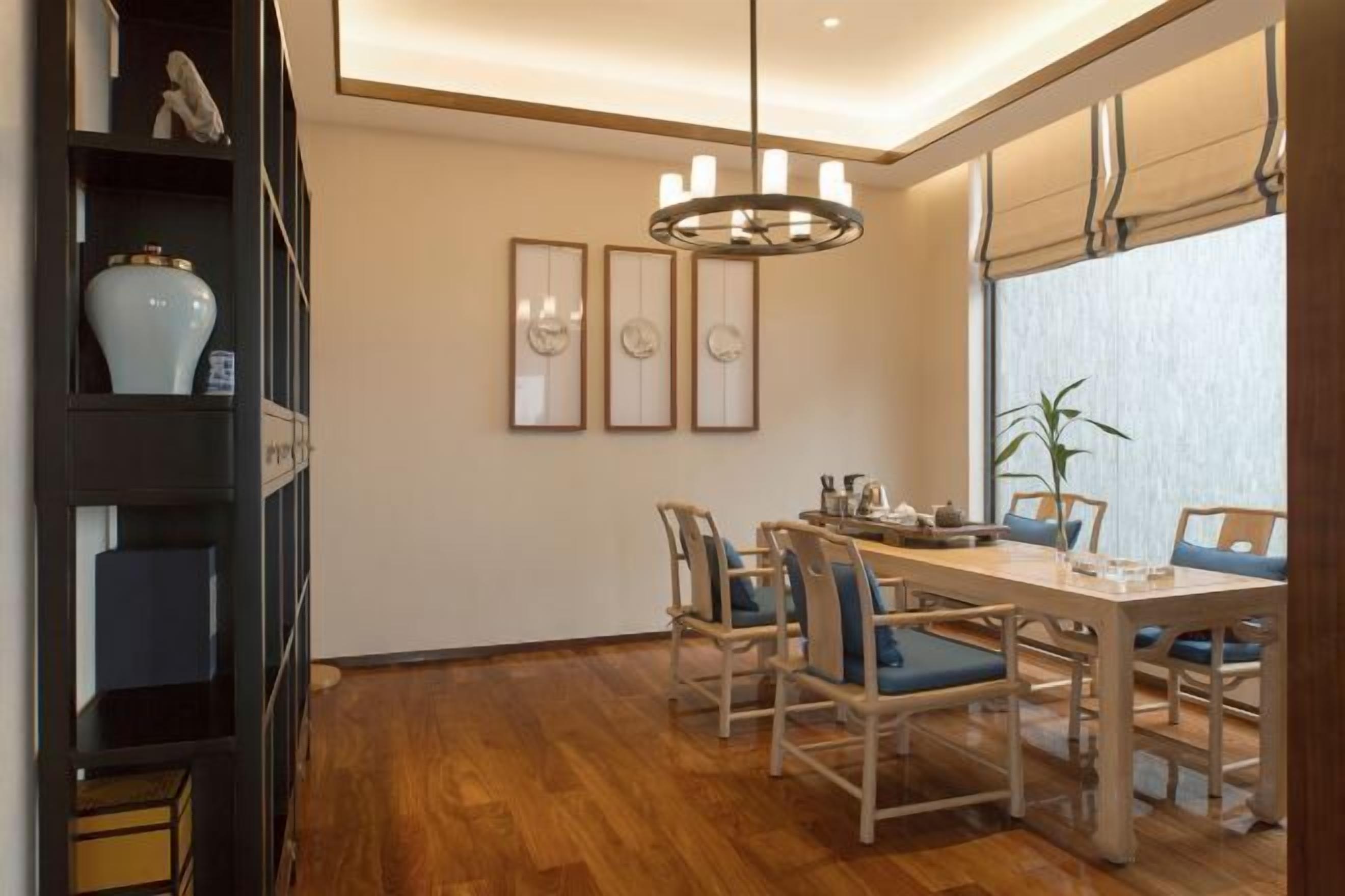A dining area with a wooden table and chairs, a chandelier, framed artwork on the wall, and a potted plant by the window.