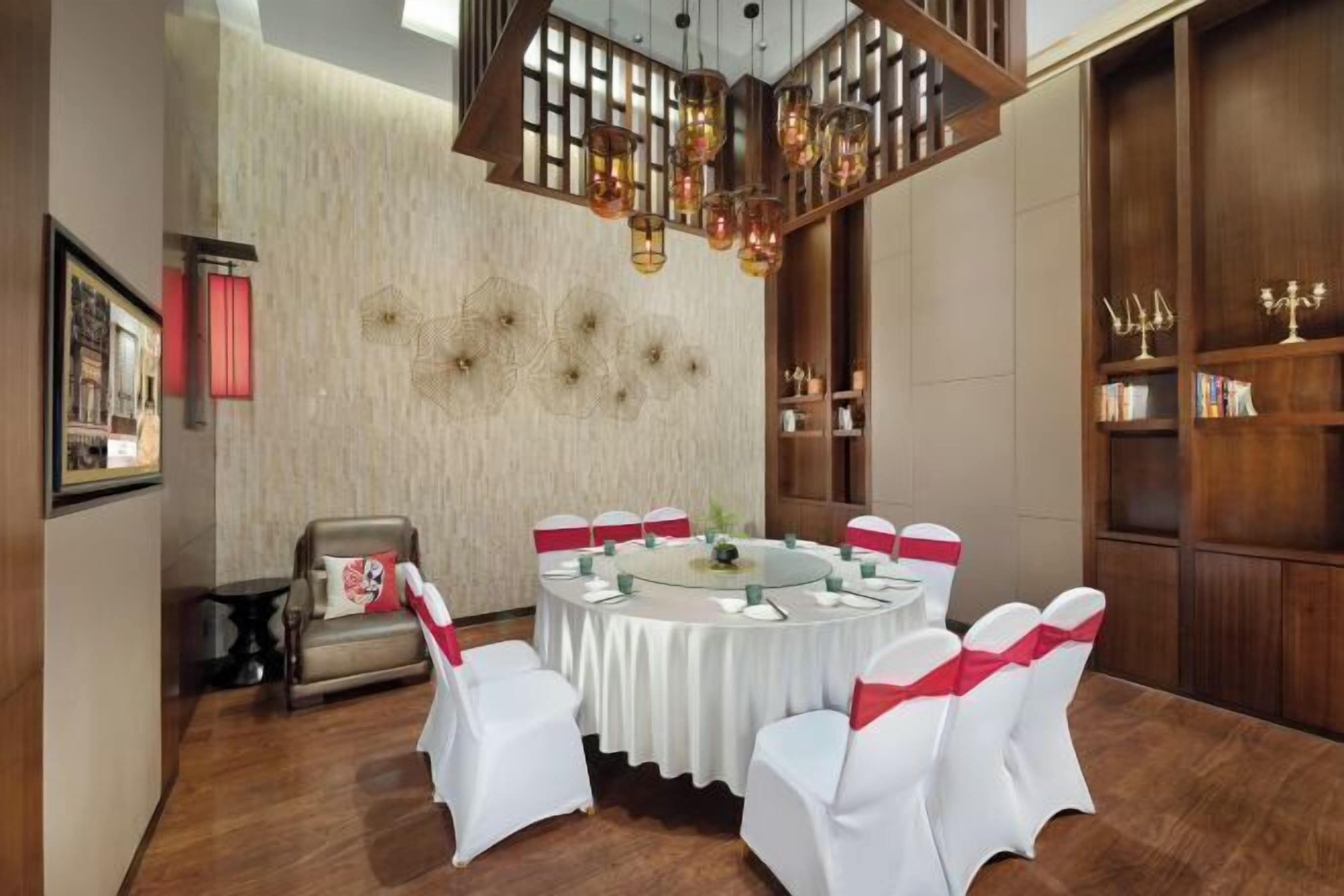 A round table with white chairs and red sashes, set in a room with wooden flooring and a decorative ceiling light fixture.