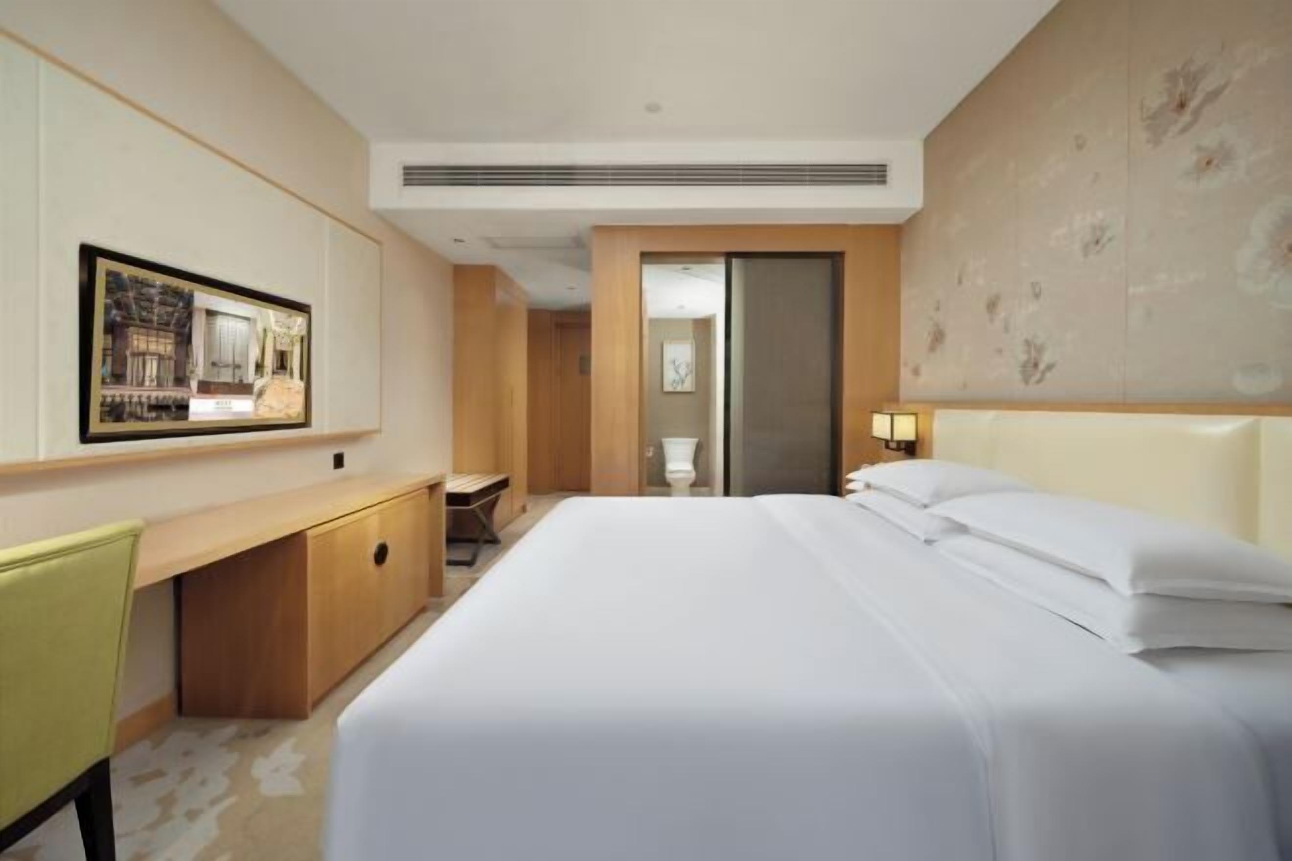 A hotel room with a large bed, a desk with a chair, a mirror, and a wall-mounted picture.