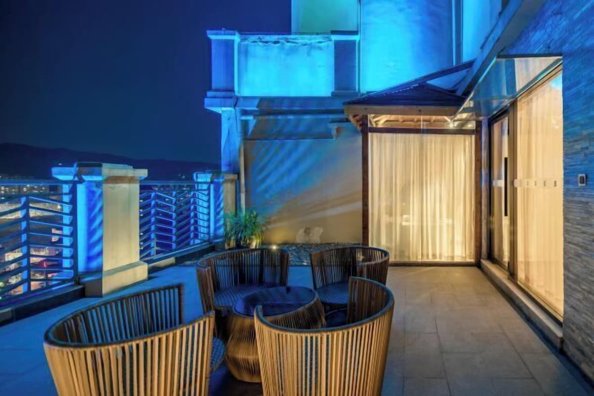 A rooftop terrace with wicker furniture, a view of the city, and a glass door leading to the interior.