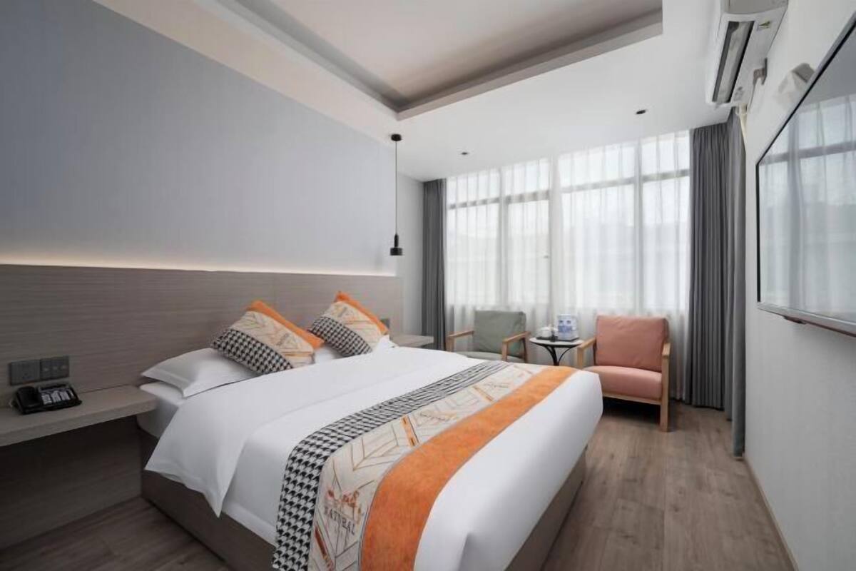 A hotel room with a large bed, a bedside table with a phone, a chair, a small table with a lamp, and a large window with curtains.