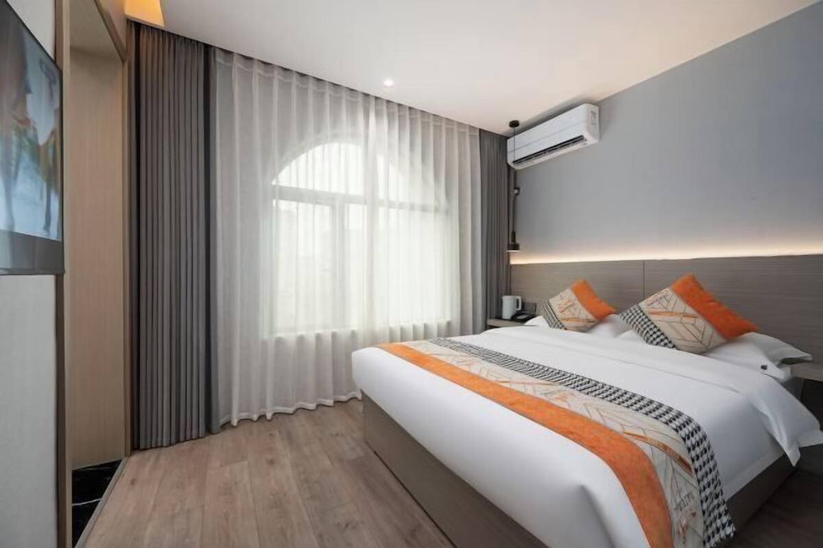 A modern hotel room with a large bed, a flat-screen TV, an air conditioning unit, and a large window with sheer curtains.