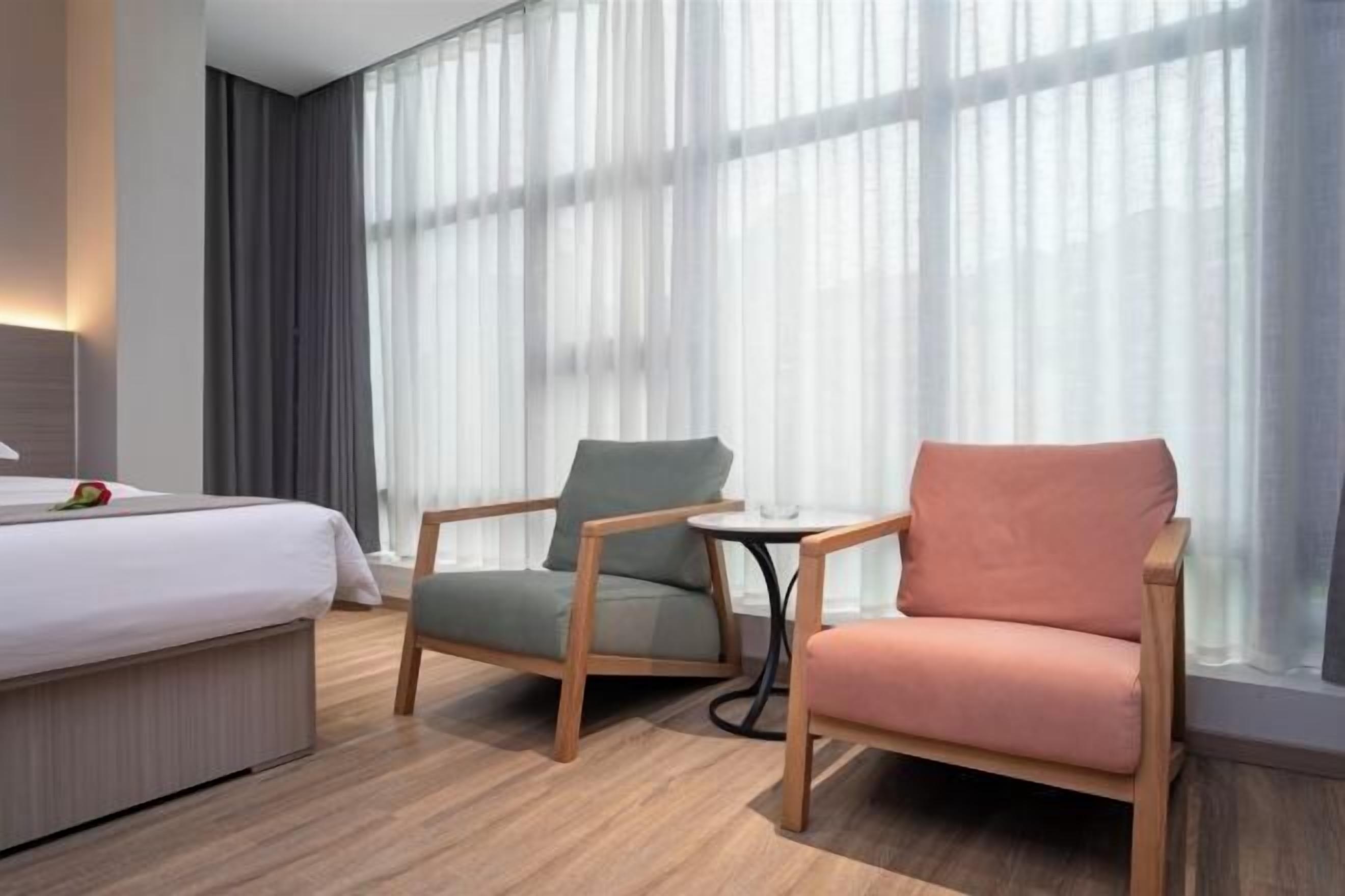 Een hotelkamer met een bed, twee stoelen, een klein tafeltje en grote ramen met dunne gordijnen.