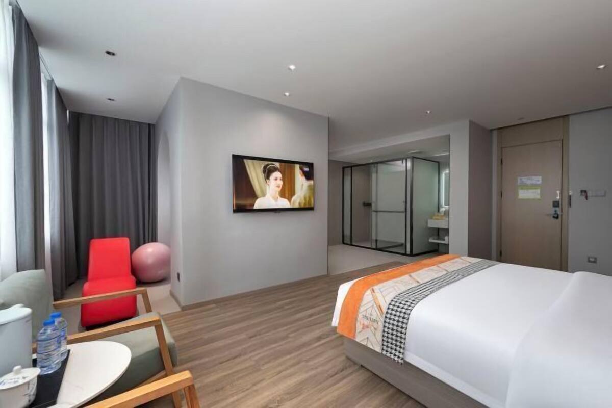 A modern hotel room with a large bed, a red chair, a glass table, and a TV on the wall.
