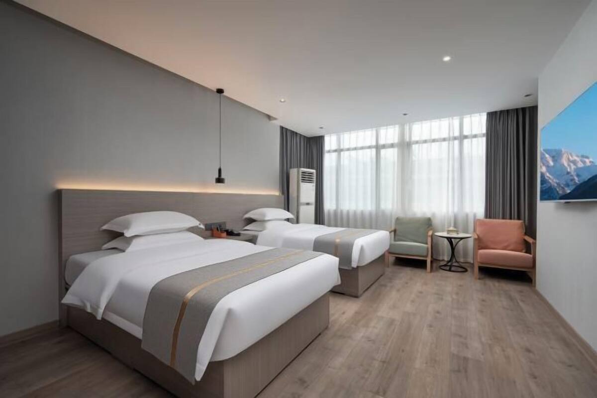 A hotel room with two beds, a small table, a chair, a TV, and a large window.