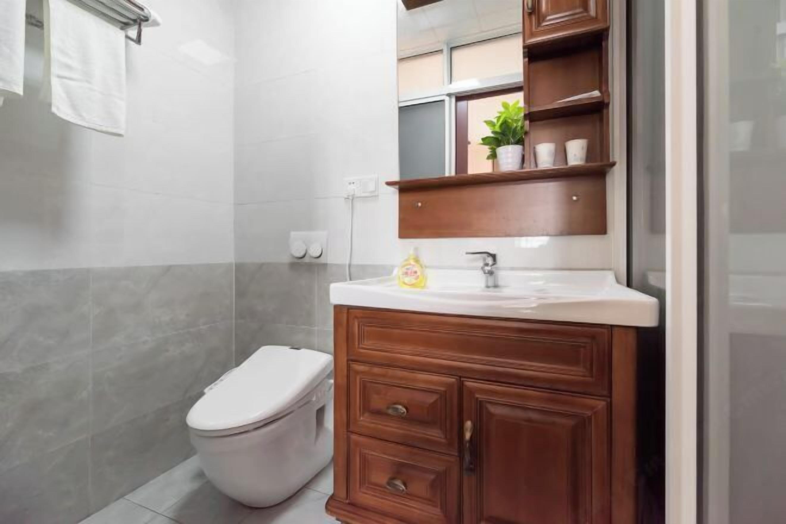 A bathroom with a white toilet, a wooden vanity with a mirror, and a shelf with a plant and cups.