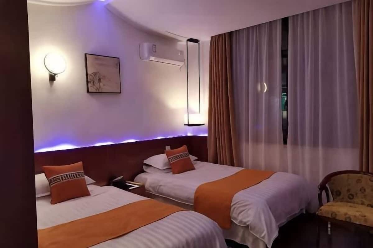 Et hotelværelse med to senge, en stol, et natbord, en lampe på væggen og et vindue med gardiner.