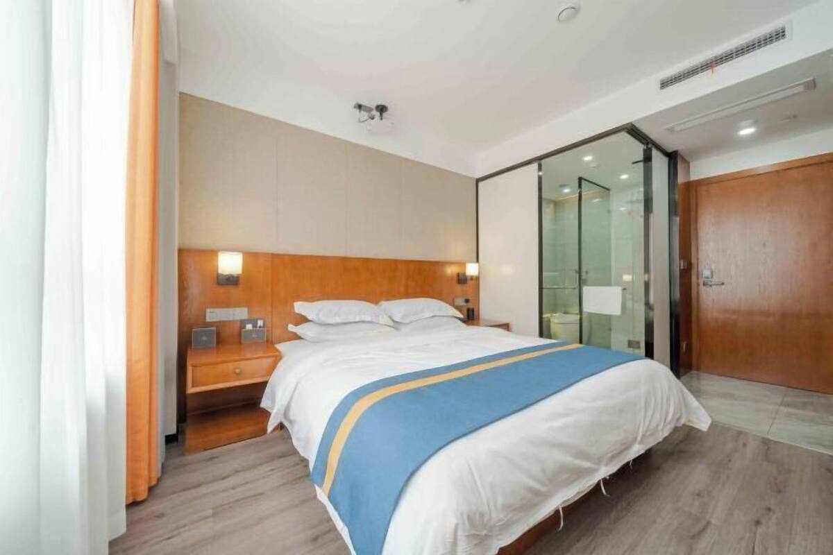 A modern hotel room with a wooden headboard, a large bed with a blue and white bedspread, a bedside table with a lamp, and a sliding glass door leading to a bathroom.