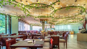 Een ruim restaurant met meerdere tafels en stoelen, een decoratief plafond met hangende lampen en planten, en grote ramen die natuurlijk licht binnenlaten.