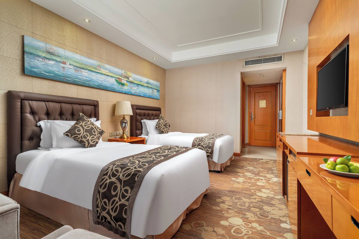 A hotel room with two beds, a TV, a painting, and a fruit plate on the desk.