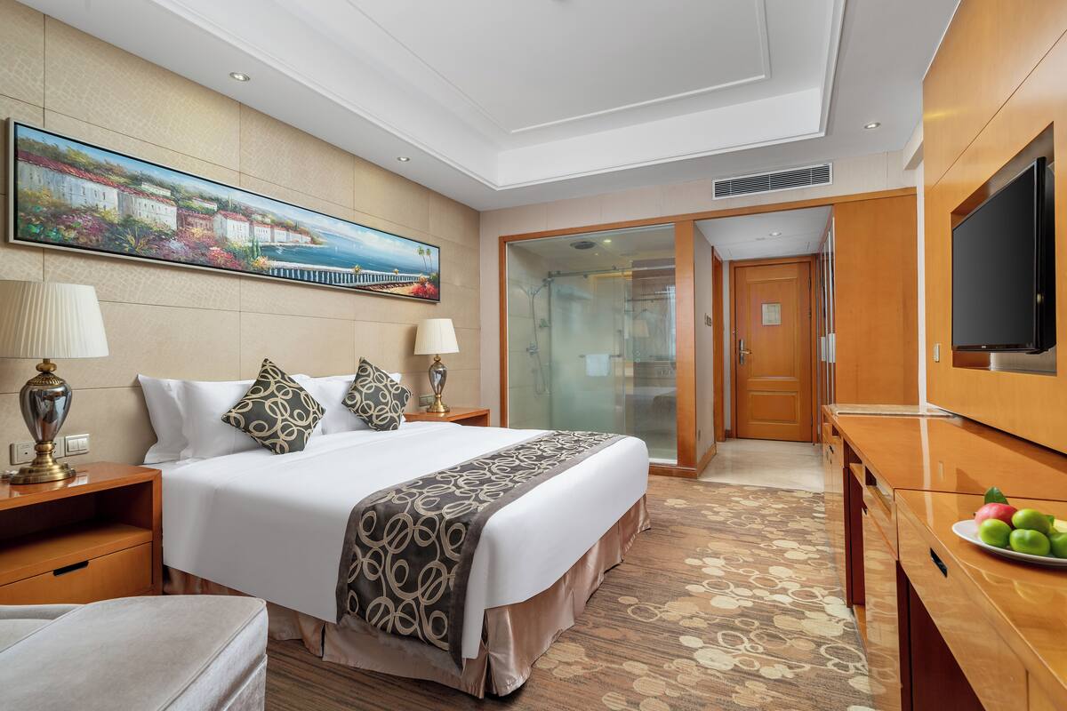 A hotel room with a large bed, a TV, a desk, and a painting on the wall.