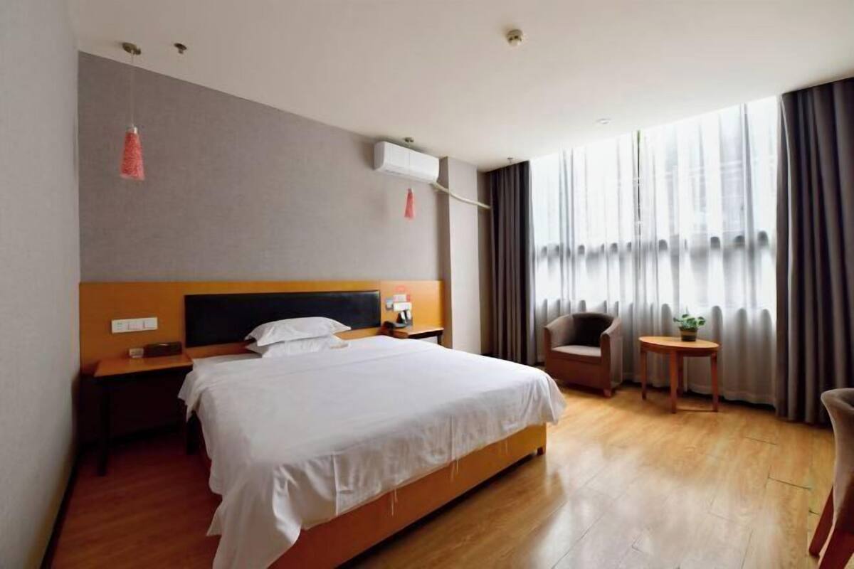 Habitació d'hotel amb un llit gran, una taula de nit, una cadira, una taula petita i una finestra amb cortines.