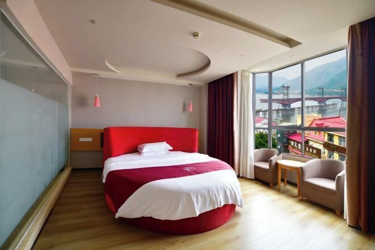 Ein Hotelzimmer mit einem großen Bett, einem kleinen Tisch, einem Sessel und Blick auf eine Brücke und Berge.