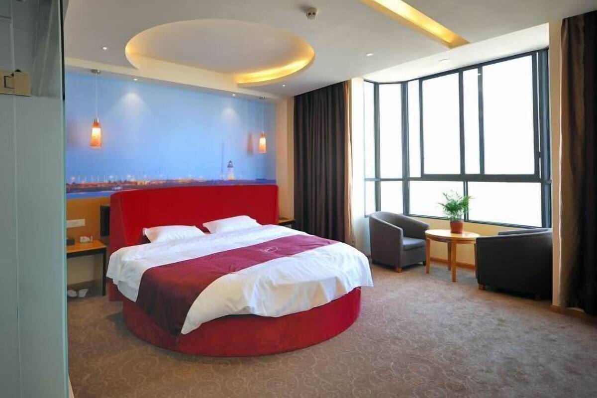Habitació d'hotel amb un llit gran, una taula de treball, una cadira i vista a un paisatge urbà a través de grans finestres.