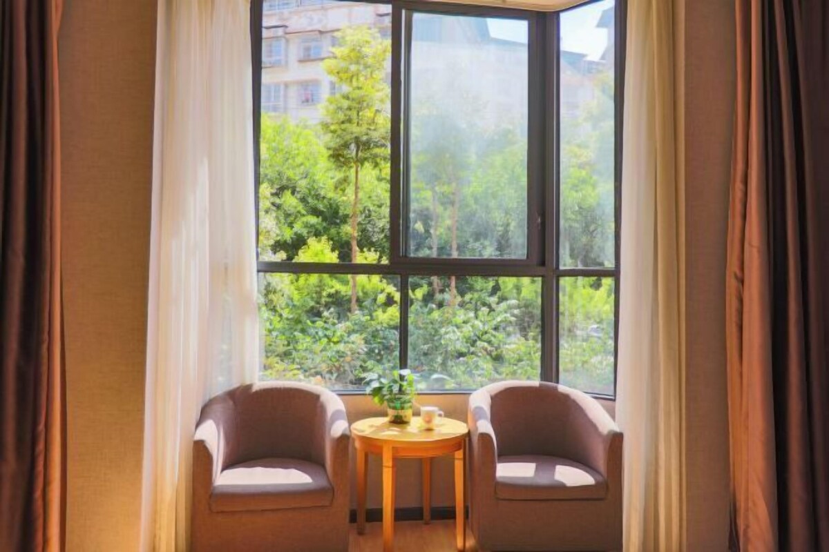 Ein Zimmer mit zwei Stühlen und einem kleinen Tisch, ein großes Fenster mit Blick auf Grünflächen und Gebäude, sowie Vorhänge am Fenster.