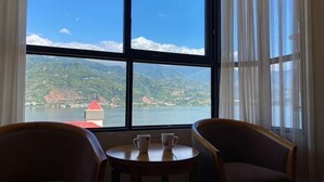 Habitación con un ventanal grande que ofrece vistas a un lago y montañas, dos sillas y una mesita con dos tazas.