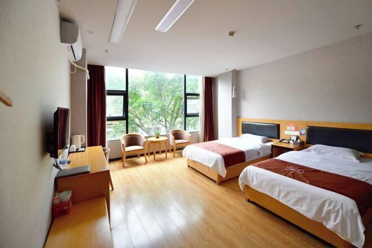 Habitació d'hotel amb dos llits, una taula de treball, una cadira, un televisor i una finestra amb vista a arbres.