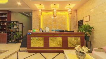 A hotel reception area with a wooden reception desk, a seating area with a vase of flowers, and a wall with framed certificates.