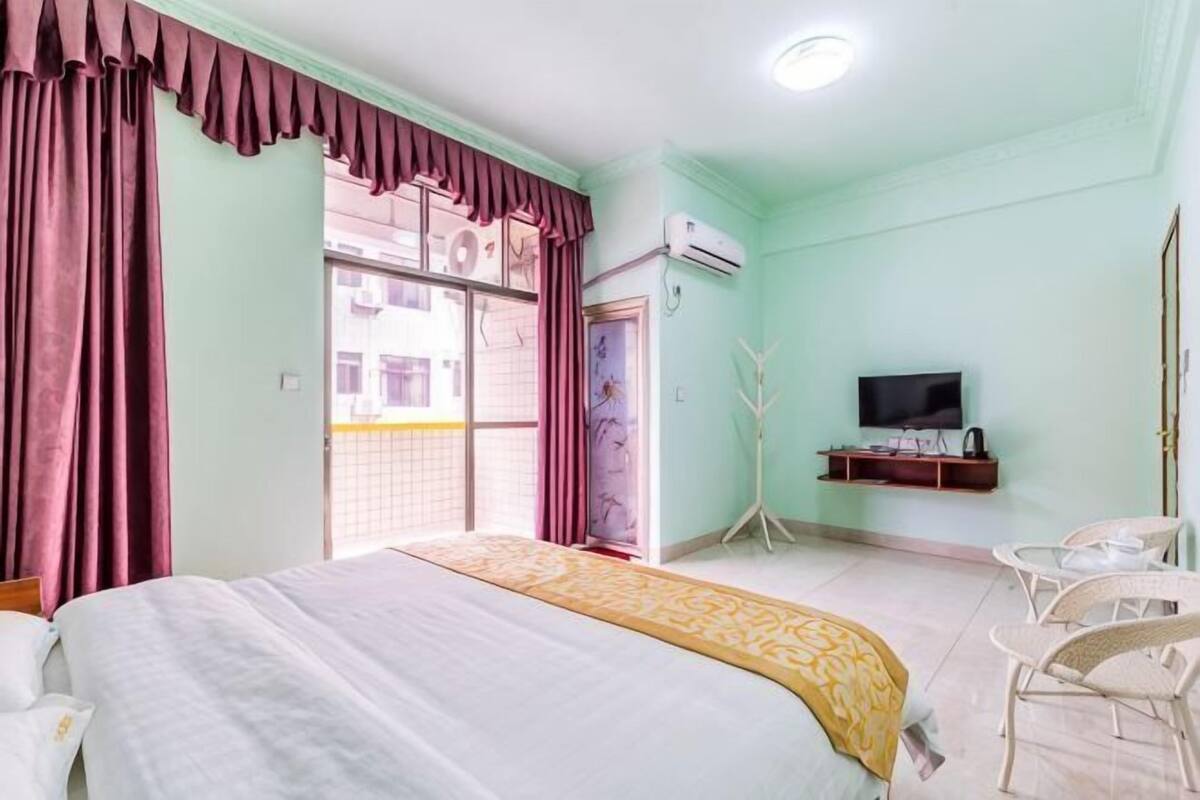 A bedroom with a bed, a TV, a chair, a window with curtains, and an air conditioning unit.