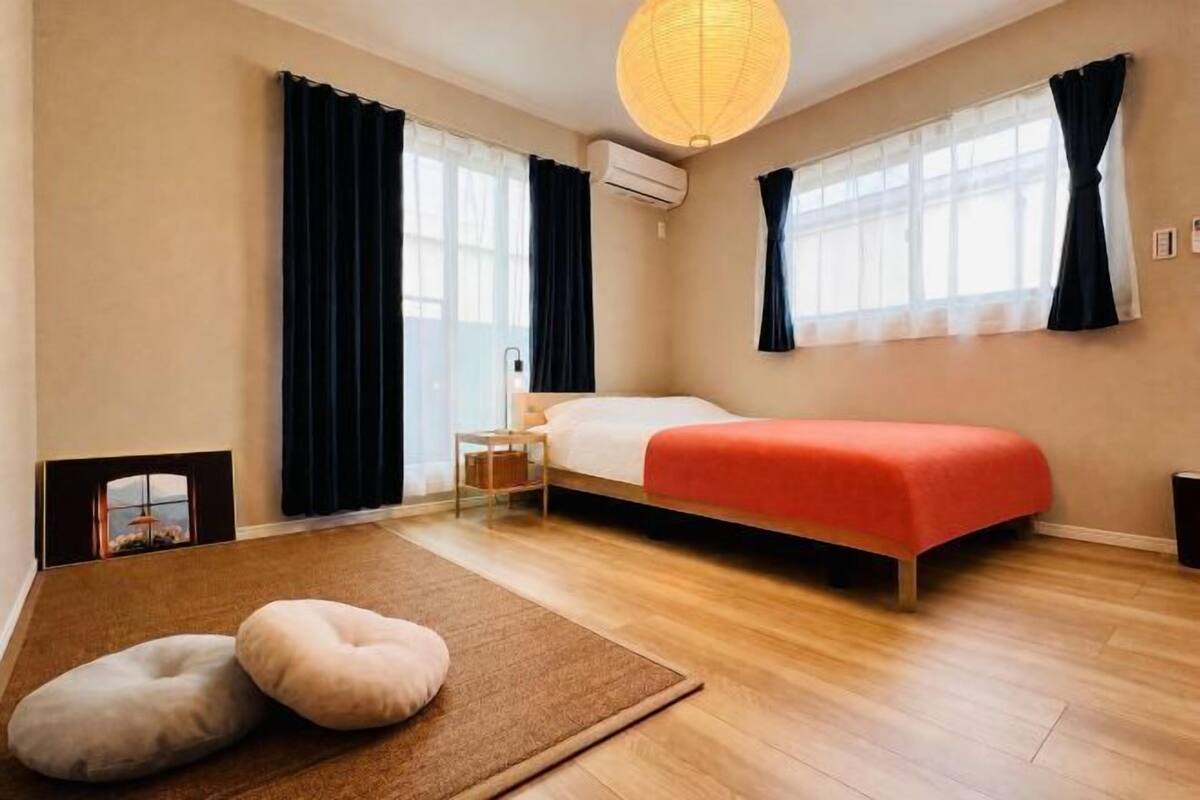 A bedroom with a bed, two windows with curtains, a wall-mounted air conditioner, a wooden floor, and a small bedside table.