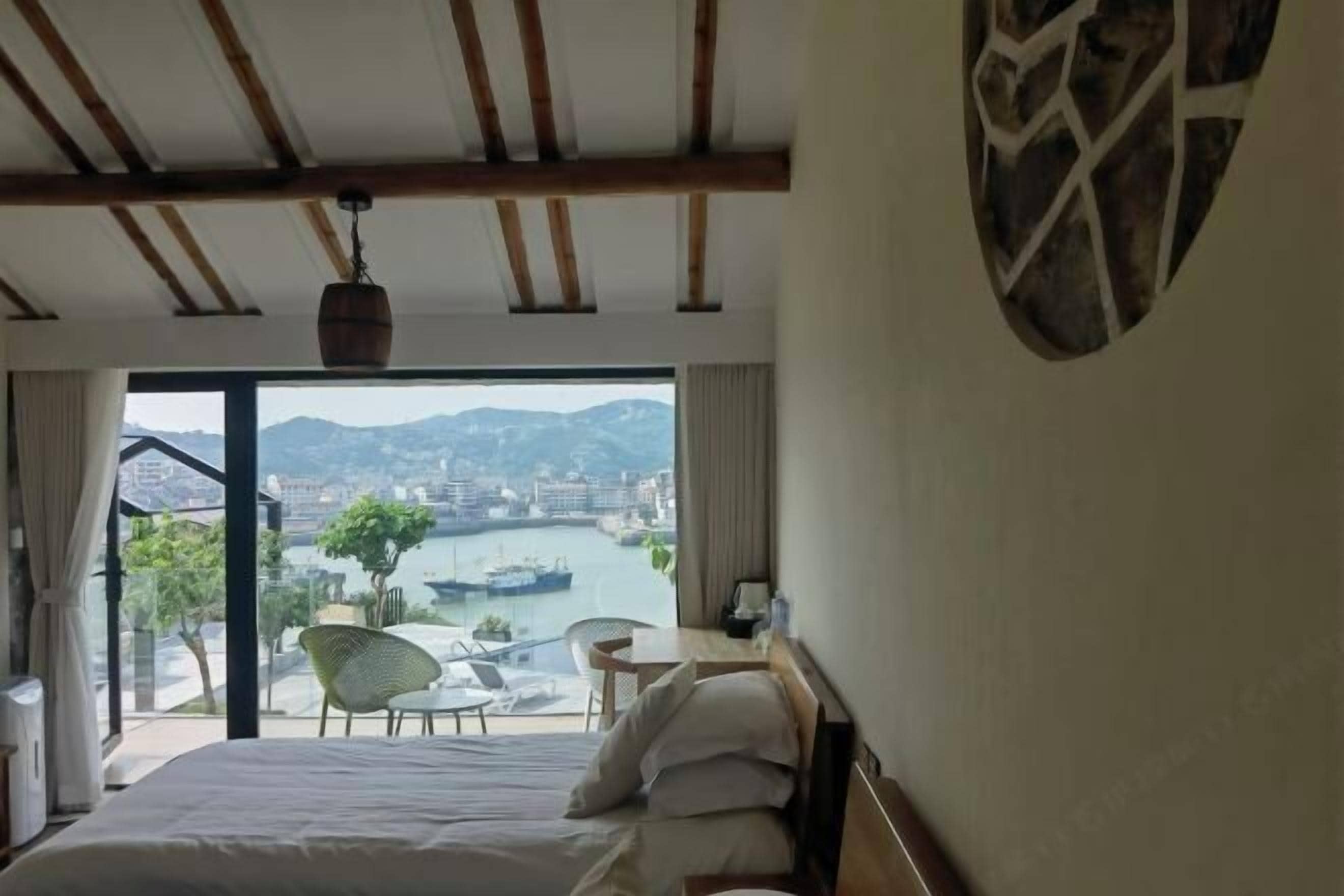 A hotel room with a bed, a view of a river and cityscape, a dining area with chairs, and a hanging light fixture.