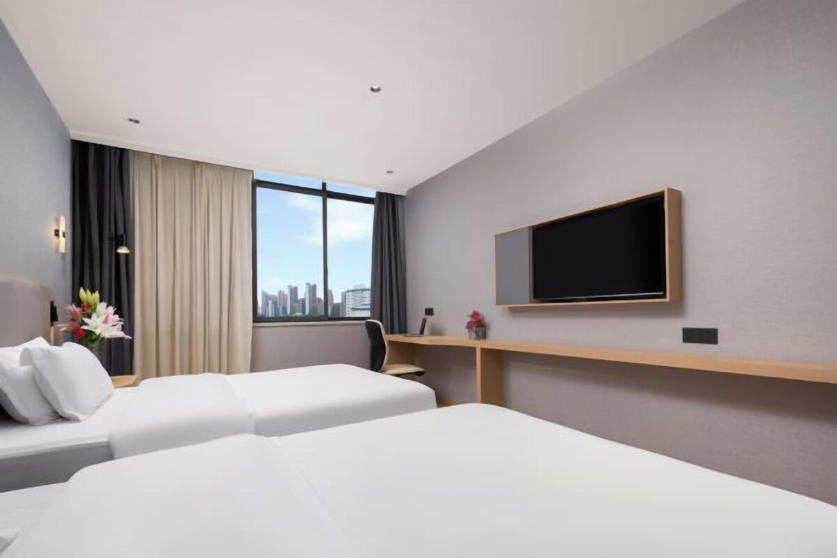 A hotel room with two beds, a TV on the wall, a desk, and a window with curtains.