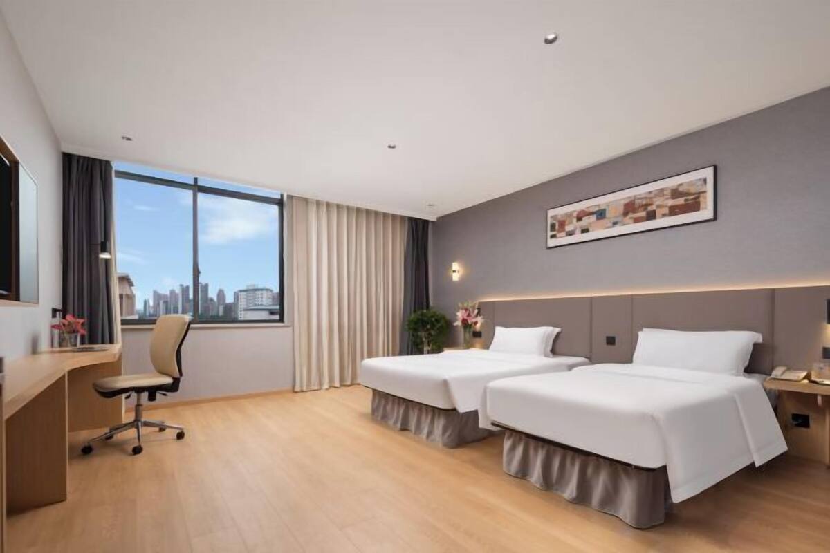 A hotel room with two beds, a desk, a chair, a window with a city view, and a framed picture on the wall.