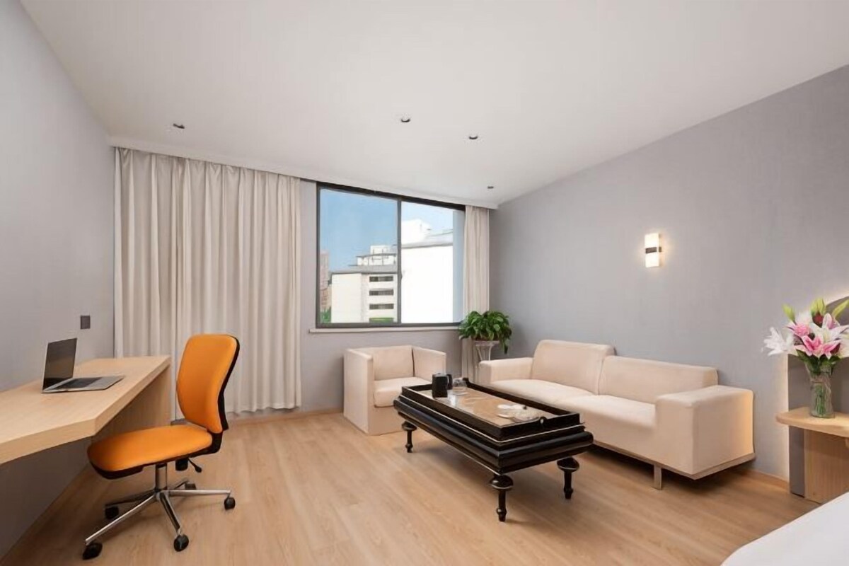 A modern hotel room with a desk, chair, sofa, coffee table, and a large window with curtains.