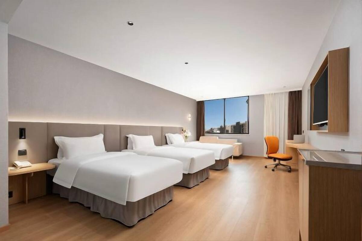 A modern hotel room with three beds, a desk, a chair, and a large window.