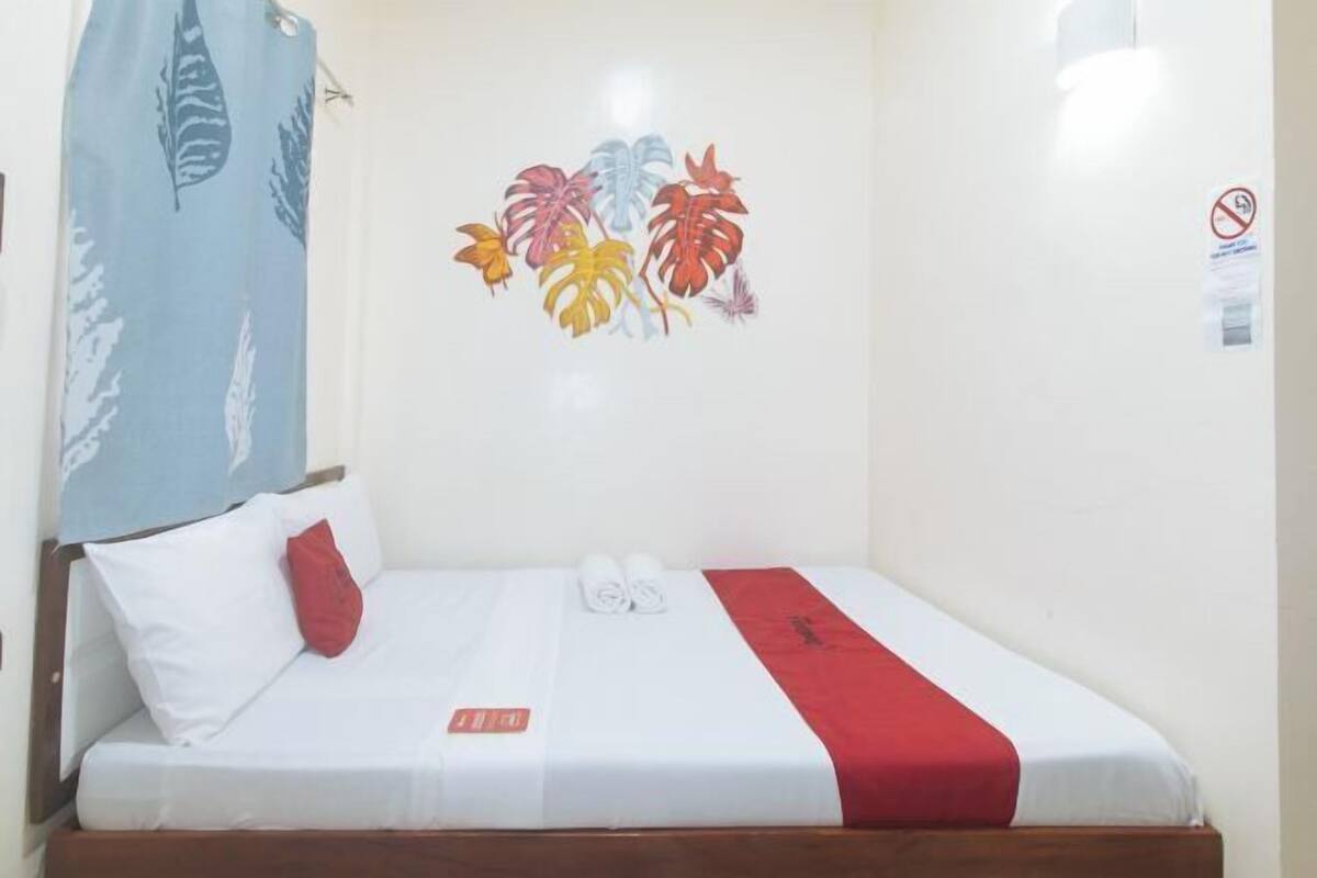 A single bed with white bedding and a red accent, a wall with a tropical leaf mural, and a no-smoking sign.
