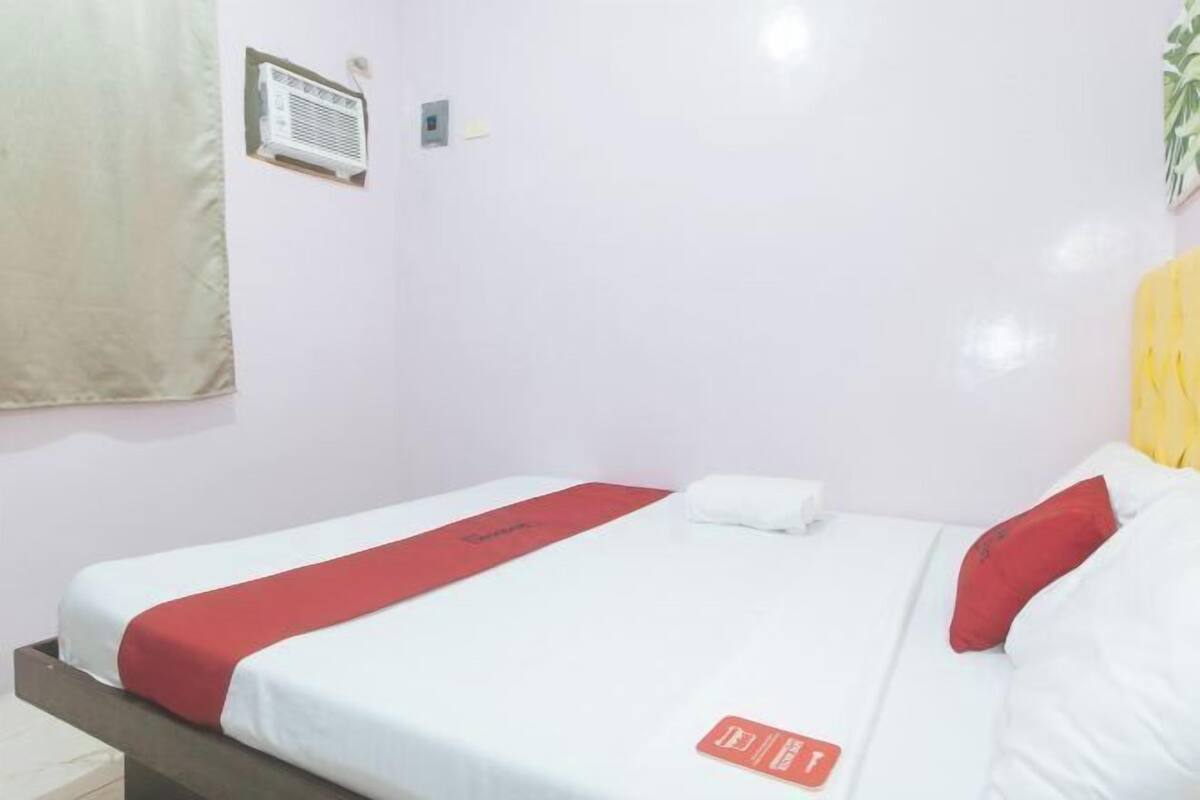 A single bed with a red and white bedspread, a white pillow, and a red pillow on a white wall with a small air conditioning unit and a light switch.